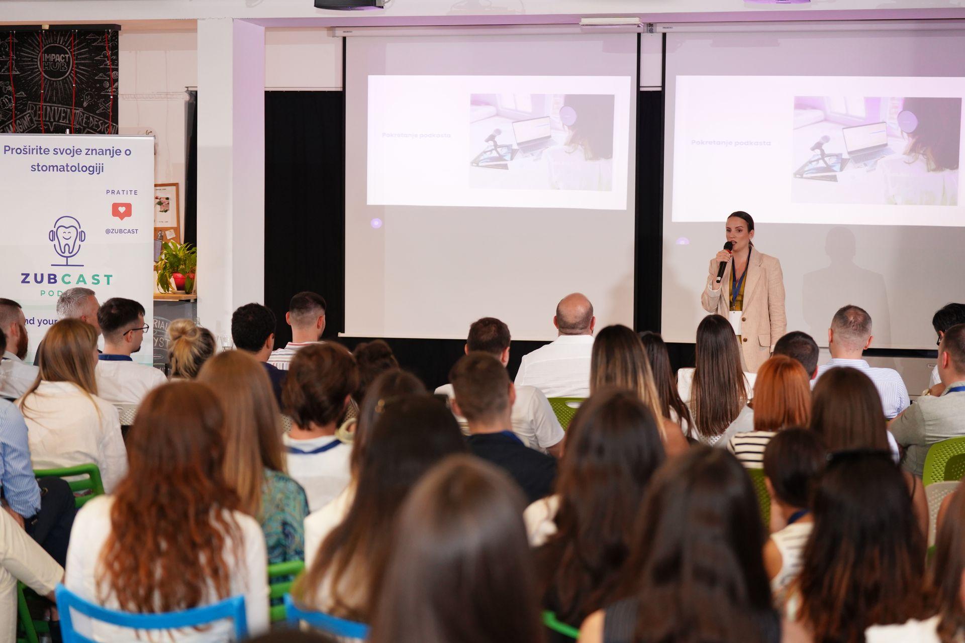 Speaker presenting in front of an audience with presentation slides on screen.
