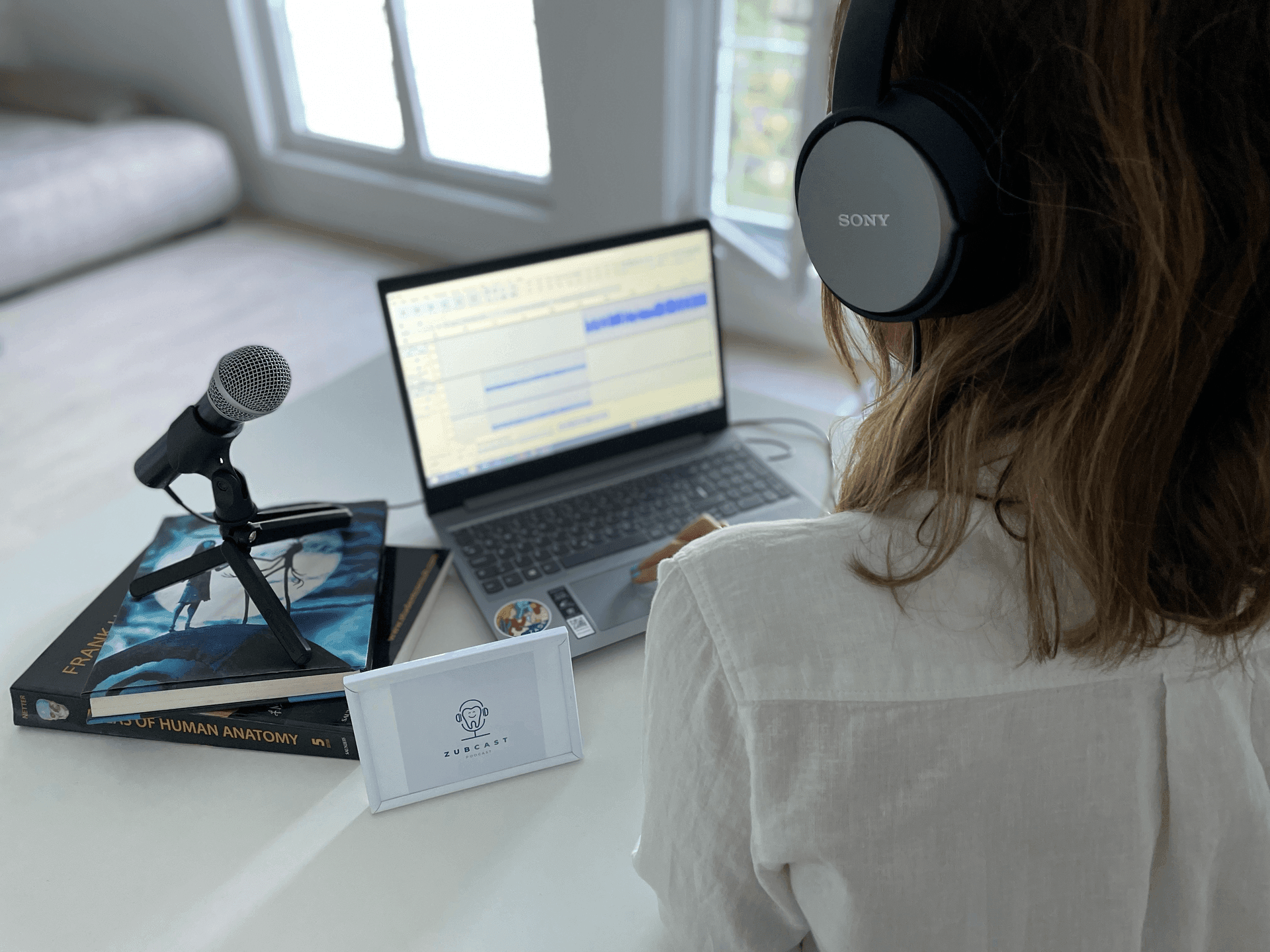 Person wearing headphones using a laptop with audio editing software, microphone on a table with anatomy books.